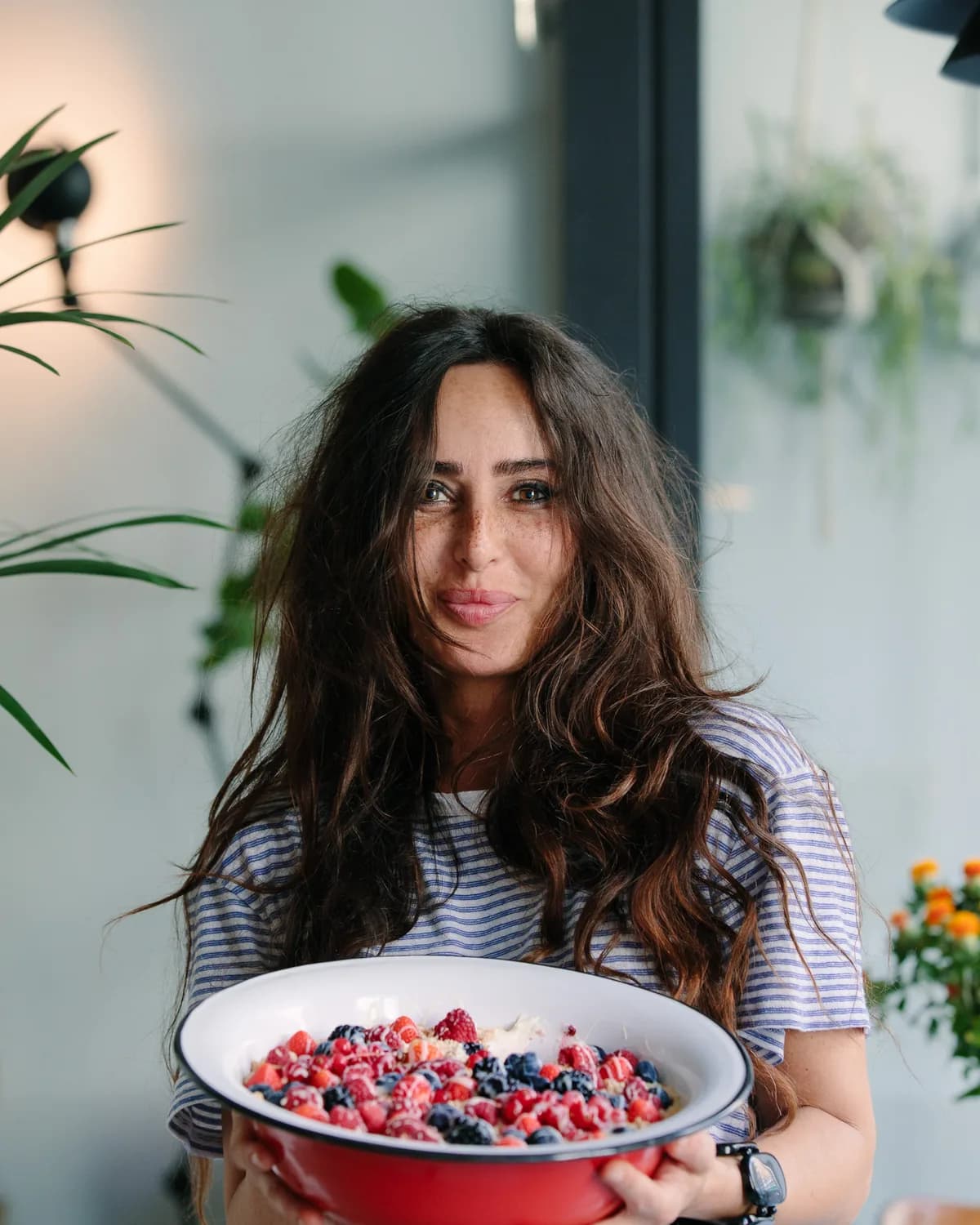 Founder holding a wide red bowl of berries and crumble in a plant-filled dining room