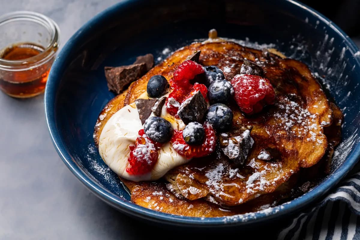 Pancakes in a deep blue bowl topped with raspberries, blueberries, cream and chocolate