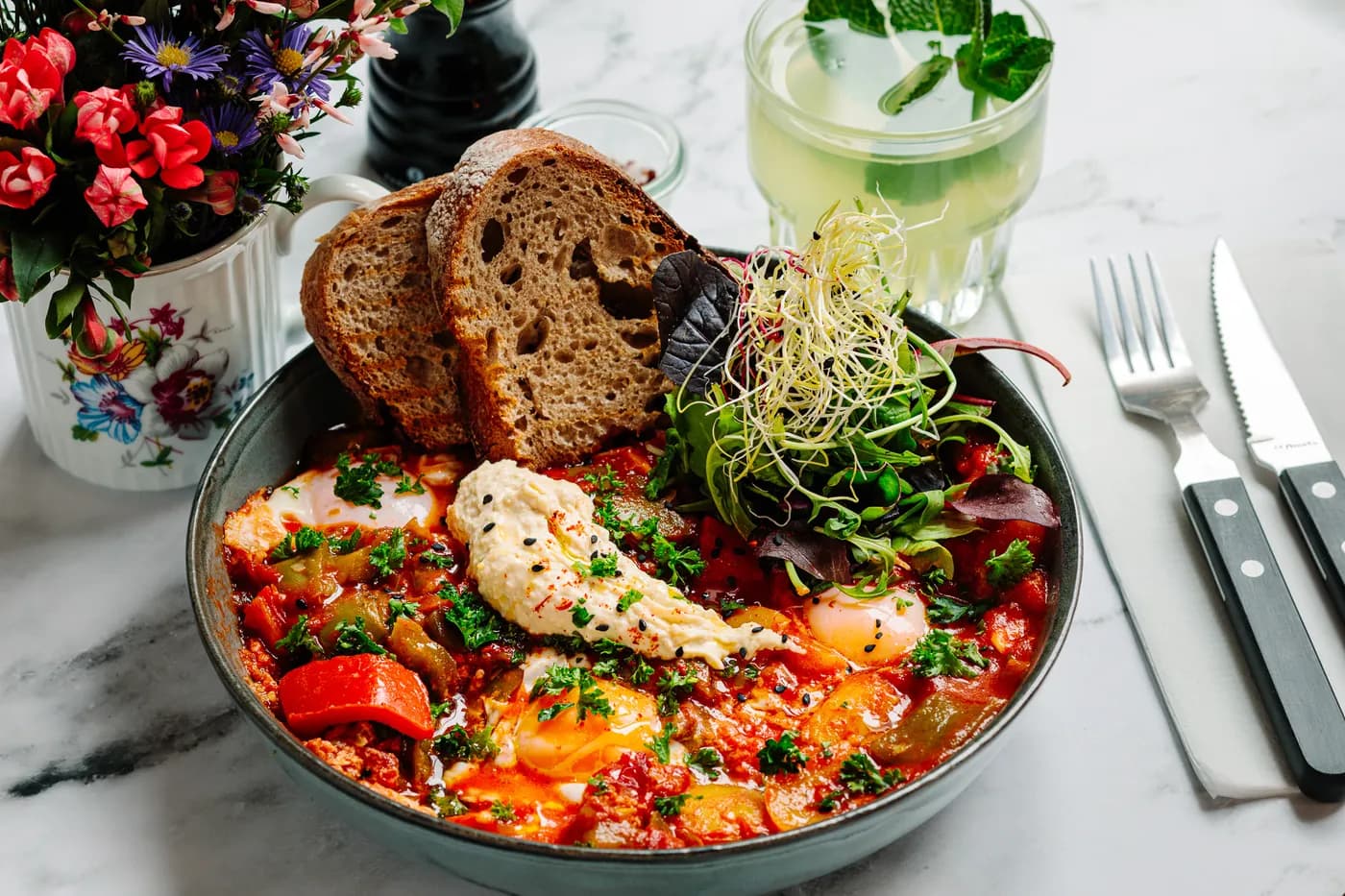 Shakshuka served in a stone bowl with sourdough, fresh greens and a cold drink